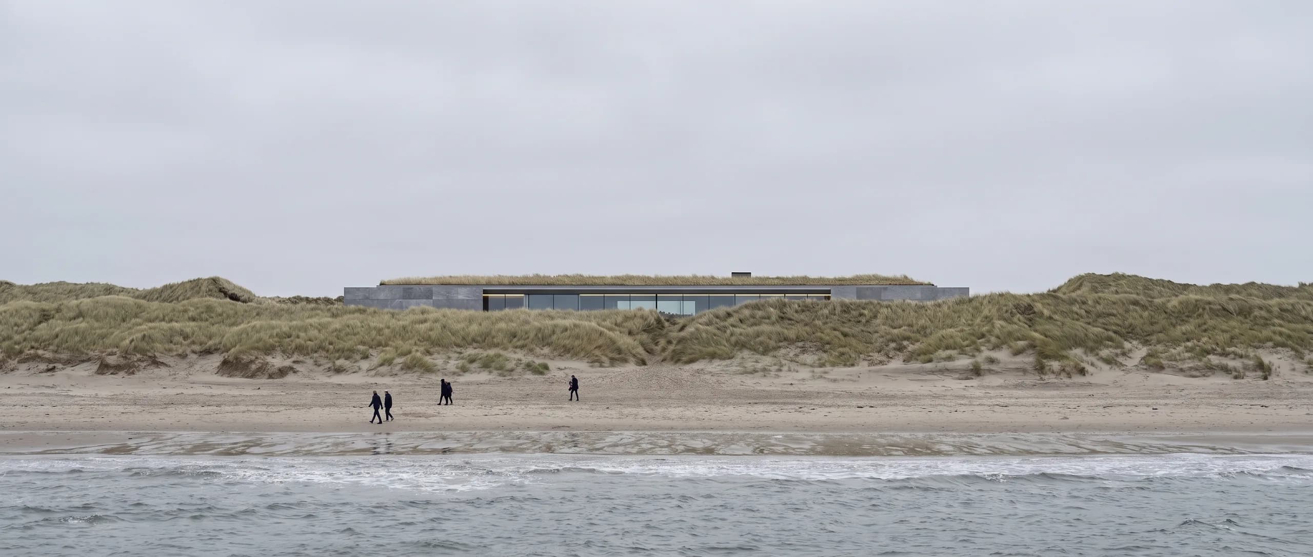 Nøytral konseptillustrasjon i dagslys sett fra den offentlige stranden i flatt, overskyet lys, som viser den lave, gresskledde bygningen bak og under den naturlige dyneryggen.